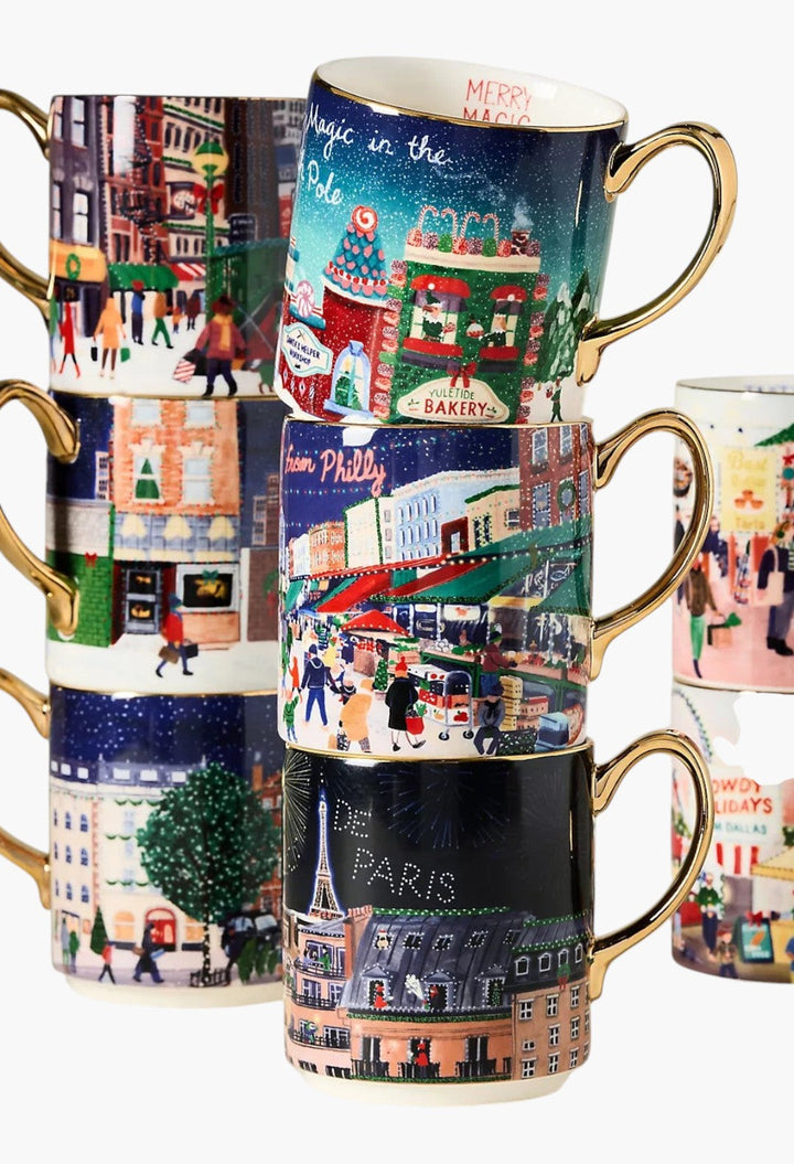 GLAM MODA Holiday in the City stoneware mugs stacked with festive streetscape illustrations and gold-painted handles.