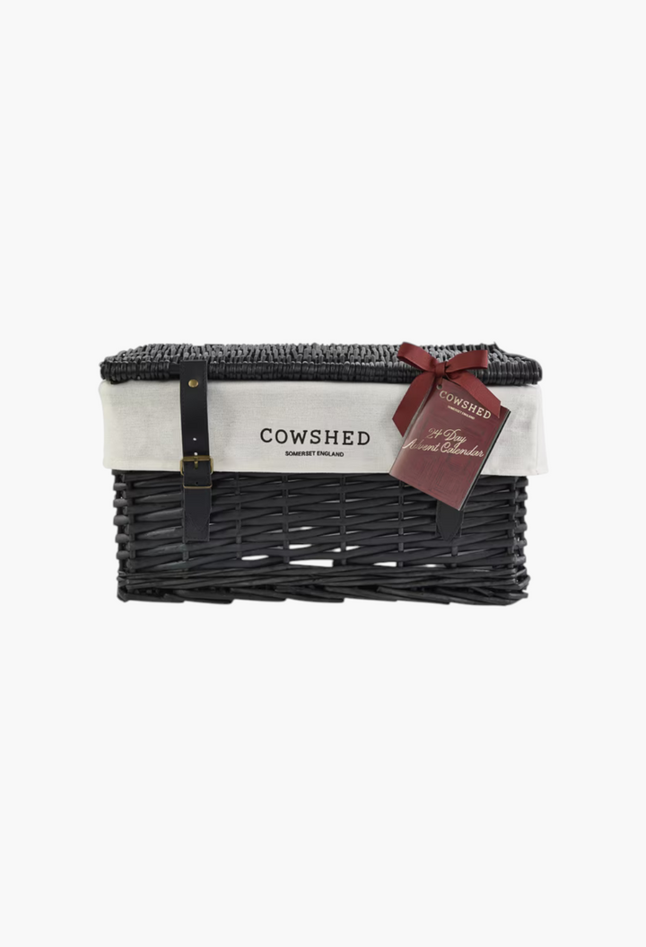 GLAM MODA Cowshed 24 Day Advent Calendar in a black handwoven wicker hamper with a white fabric lining and red ribbon.