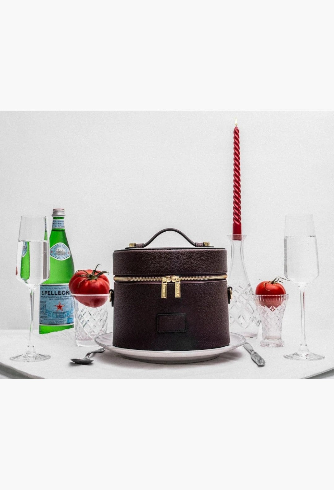 GLAM MODA oval vegan leather toiletry case in dark brown with gold dual zippers, presented on a dining table setting.