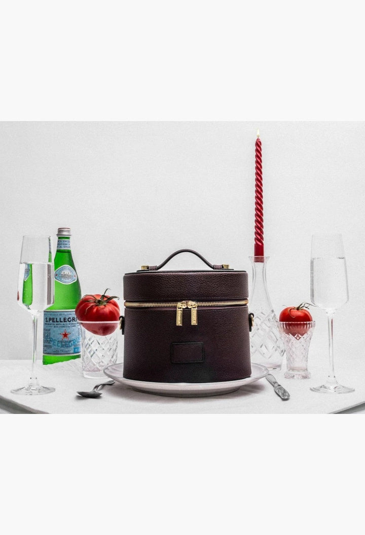 GLAM MODA oval vegan leather toiletry case in dark brown with gold dual zippers, presented on a dining table setting.