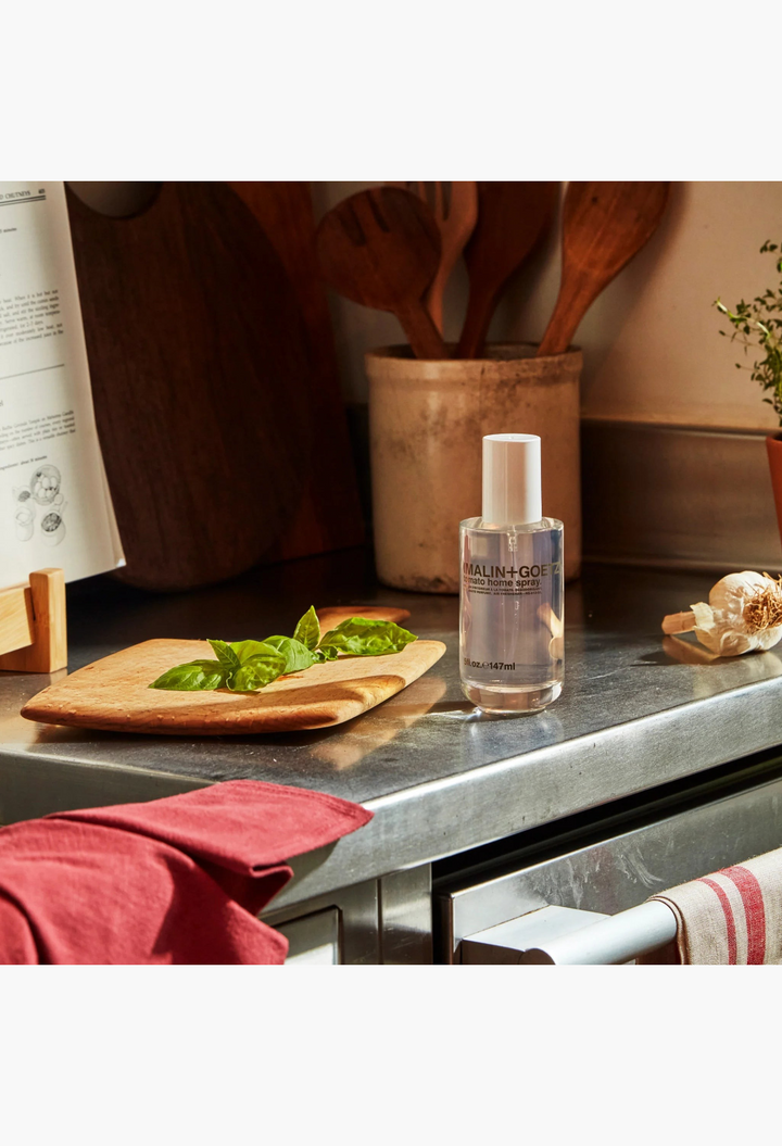 GLAM MODA Malin+Goetz tomato home spray bottle on a kitchen counter with fresh basil and cooking utensils.