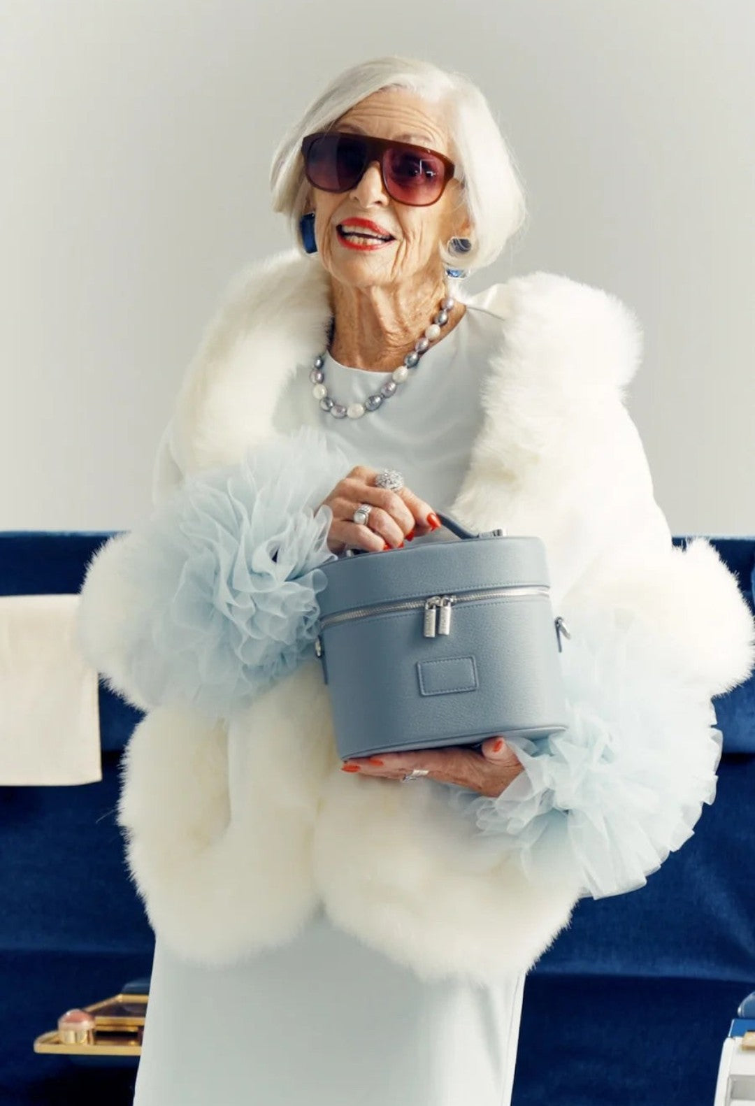 GLAM MODA oval vegan leather toiletry case in light blue held by an elegant woman in a white fur coat and sunglasses.