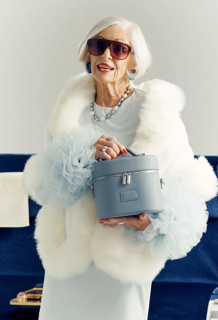 GLAM MODA oval vegan leather toiletry case in light blue held by an elegant woman in a white fur coat and sunglasses.
