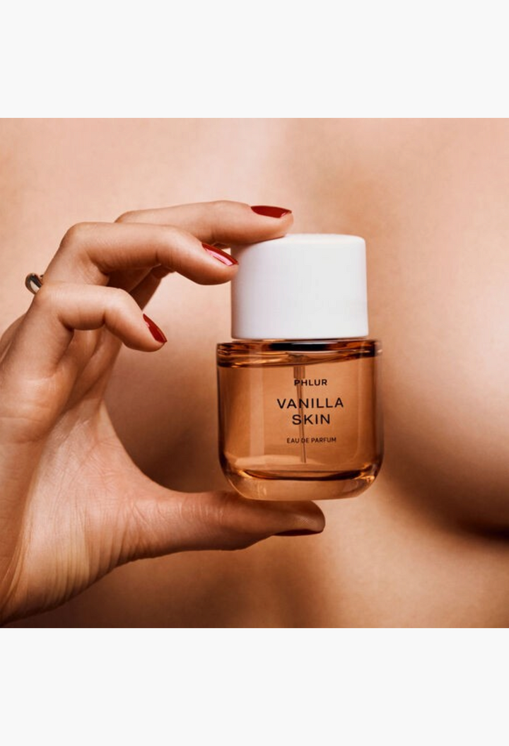 GLAM MODA Phlur Vanilla Skin Eau De Parfum bottle held by a hand with red nail polish against a warm skin background.