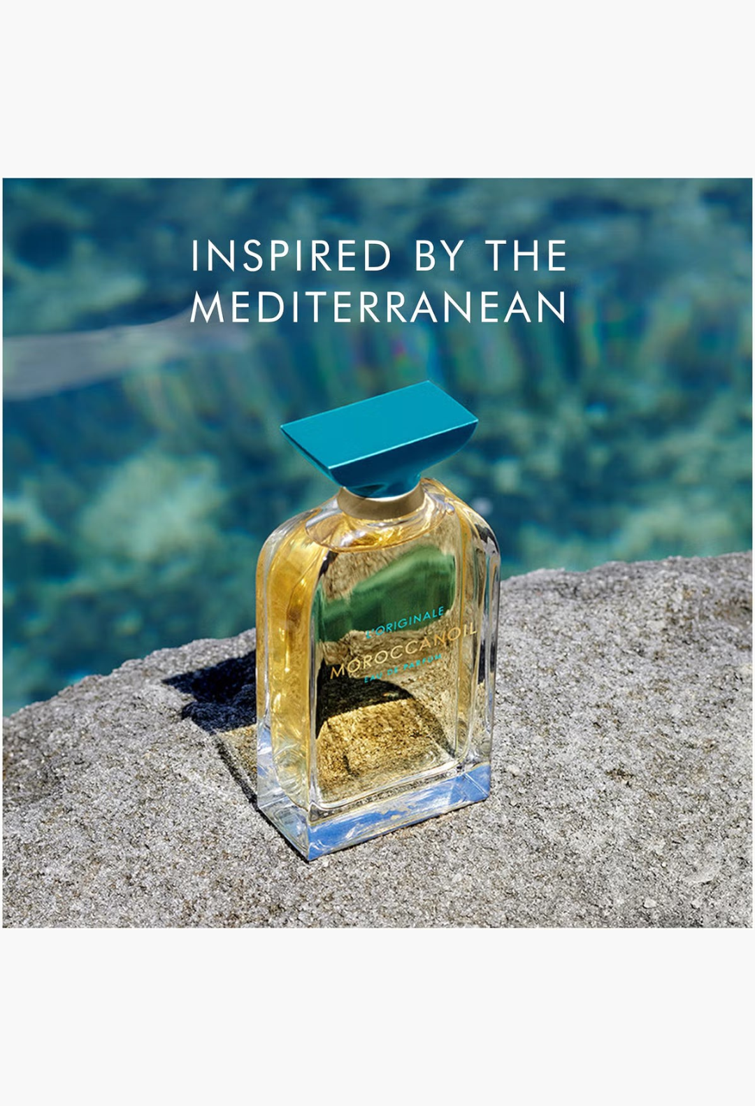 GLAM MODA Moroccanoil L'Originale Eau de Parfum bottle with a turquoise cap resting on a stone by blue water