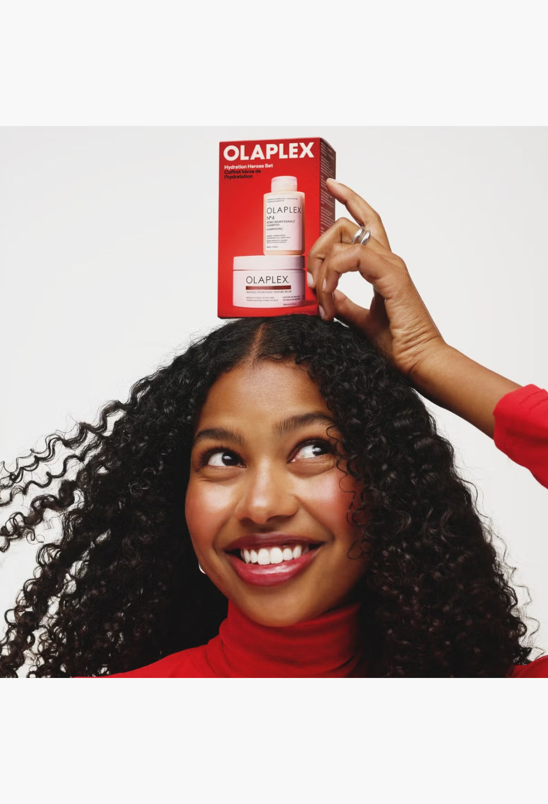 GLAM MODA model with curly hair balancing an OLAPLEX Hydration Heroes hair care set box on her head
