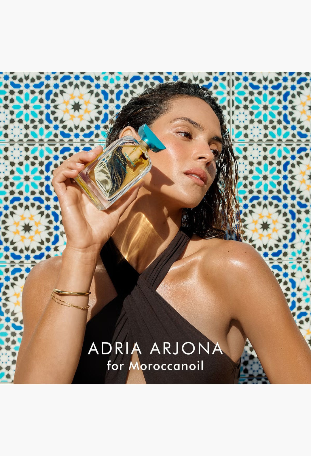GLAM MODA model Adria Arjona holding Moroccanoil L'Originale Eau de Parfum against a Mediterranean tile background.