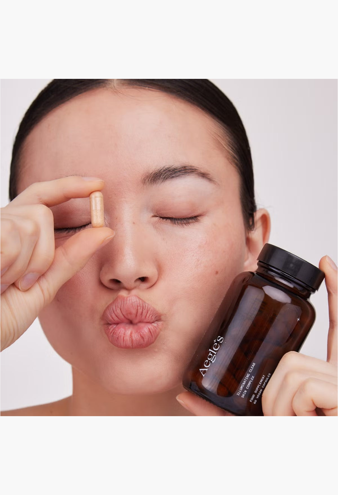 GLAM MODA model holding a capsule and a bottle of Aegle Clear Skin Complex supplement for blemish-prone skin