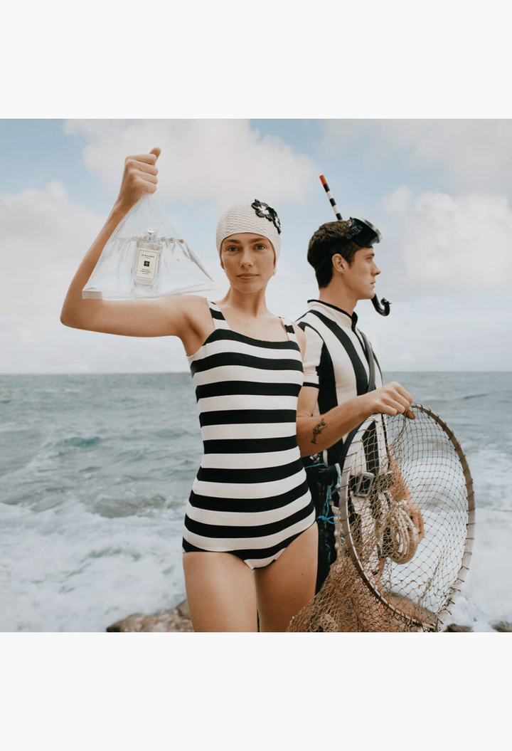 GLAM MODA Sea Salt & Bergamot Cologne held by a woman in a vintage striped swimsuit on a rocky British coastline.