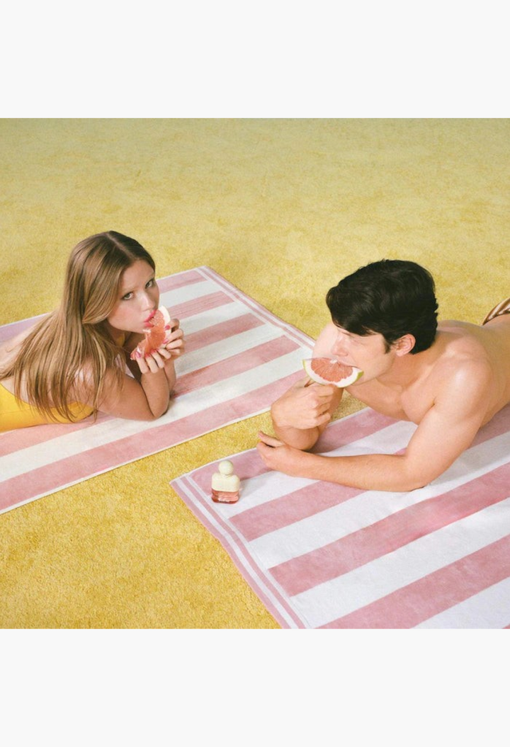 GLAM MODA Solar Drip perfume bottle on a striped beach towel with a couple eating fresh pomelo slices
